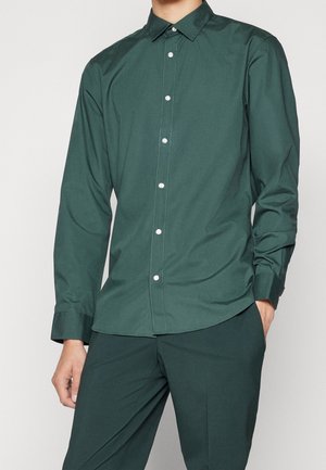 Homme portant une chemise boutonnée à manches longues vert foncé et un pantalon assorti, une main dans la poche, sur un fond uni.