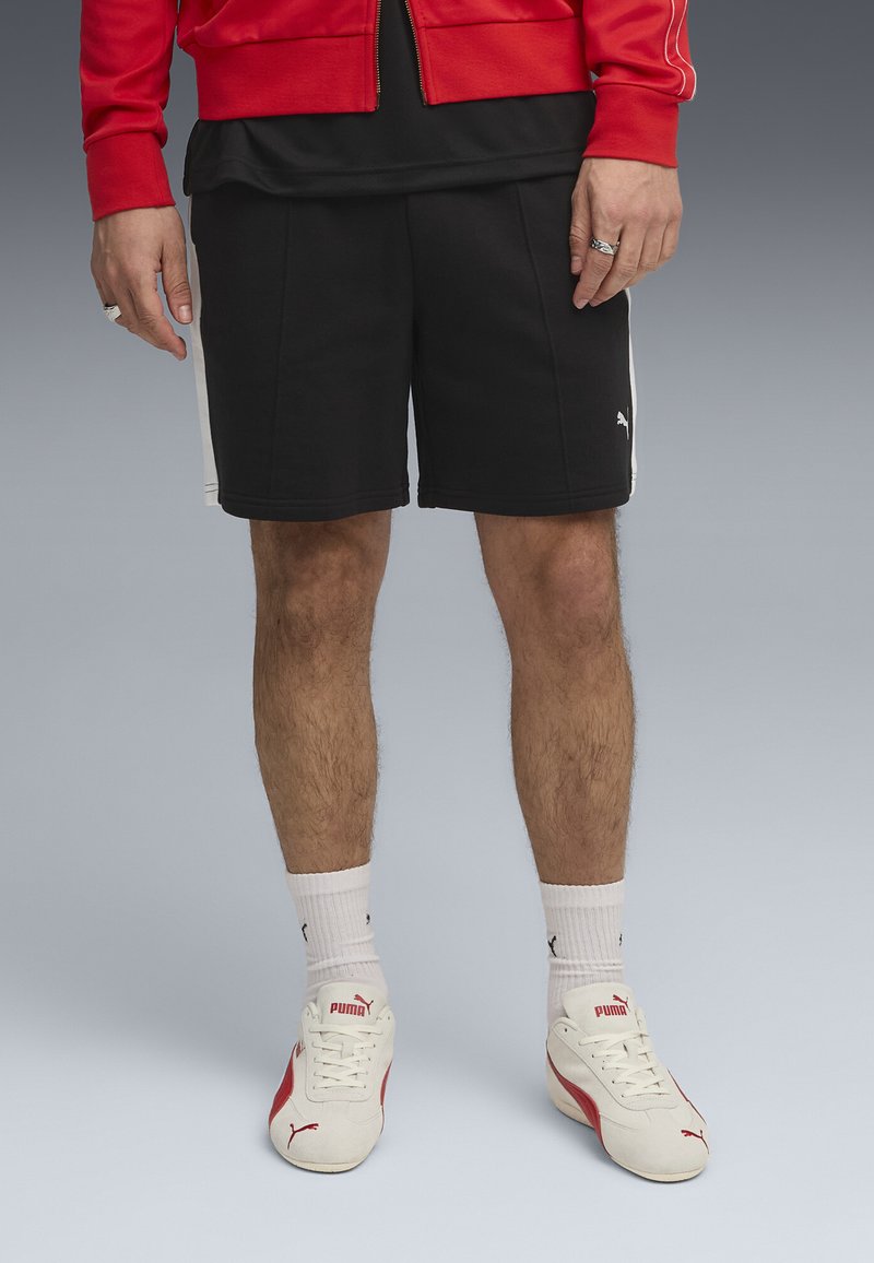 Jambes masculines portant un short noir Puma, des chaussettes blanches Puma et des baskets blanches Puma avec des accents rouges sur un fond gris.