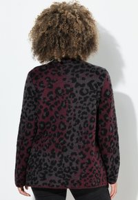 Person mit lockigem Haar, der einen langärmeligen, burgunderfarbenen und schwarzen Leopardenmuster-Pullover trägt, von hinten vor einem weißen Hintergrund betrachtet.