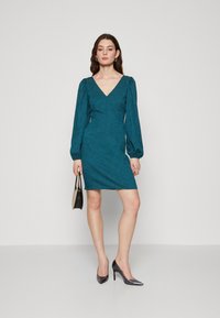 Robe teal avec un décolleté en V et des manches bouffantes, fabriquée dans un tissu texturé. Portée avec des talons hauts gris et un petit sac noir.