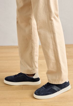 Navy suede sneakers with perforated design, white accent at the heel, and cream rubber sole. Paired with light beige pants.
