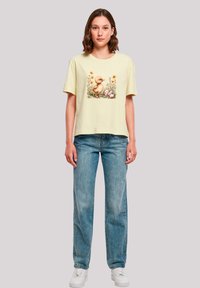 Light yellow cotton t-shirt with a front duck and flower graphic, short sleeves, and a relaxed fit. Paired with blue straight-leg jeans and white sneakers.
