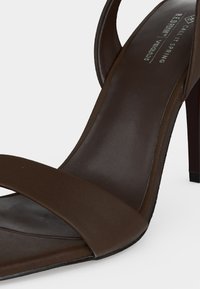 Gros plan sur une sandale marron à talon haut avec une bride avant unique et une semelle intérieure rembourrée étiquetée comme matériau végan à grain rouge.
