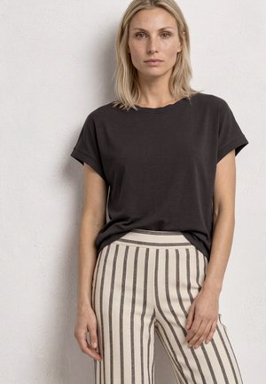 Lyshåret kvinde står op ad en tekstureret væg iført en løs sort kortærmet top og cremefarvede højtaljede bukser med lodrette sorte striber.