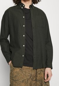 Tumma vihreä nappitakkipaita, jossa on kaulus, rintatasku ja rento istuvuus, kerroksittain mustan t-paidan päällä.