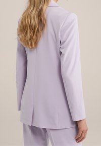 Kvinna med långt blont hår som bär en ljuslila skräddarsydd blazer och matchande byxor, sedd bakifrån mot en beige bakgrund.