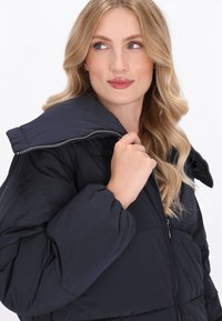 Geacă pufă navy cu guler înalt, față cu fermoar și manșete strânse. Textură netedă, matlasată și un design ușor.