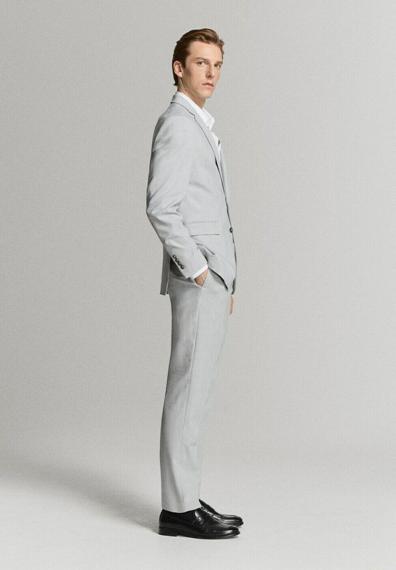 Jonge man die zijwaarts staat, gekleed in een lichtgrijs pak met zwarte schoenen, tegen een effen lichtgrijze achtergrond.