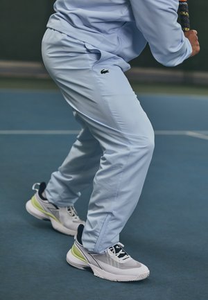 Personne en tenue de sport bleu clair et baskets blanches tenant une raquette de tennis, positionnée sur un court de tennis bleu, se préparant à jouer.