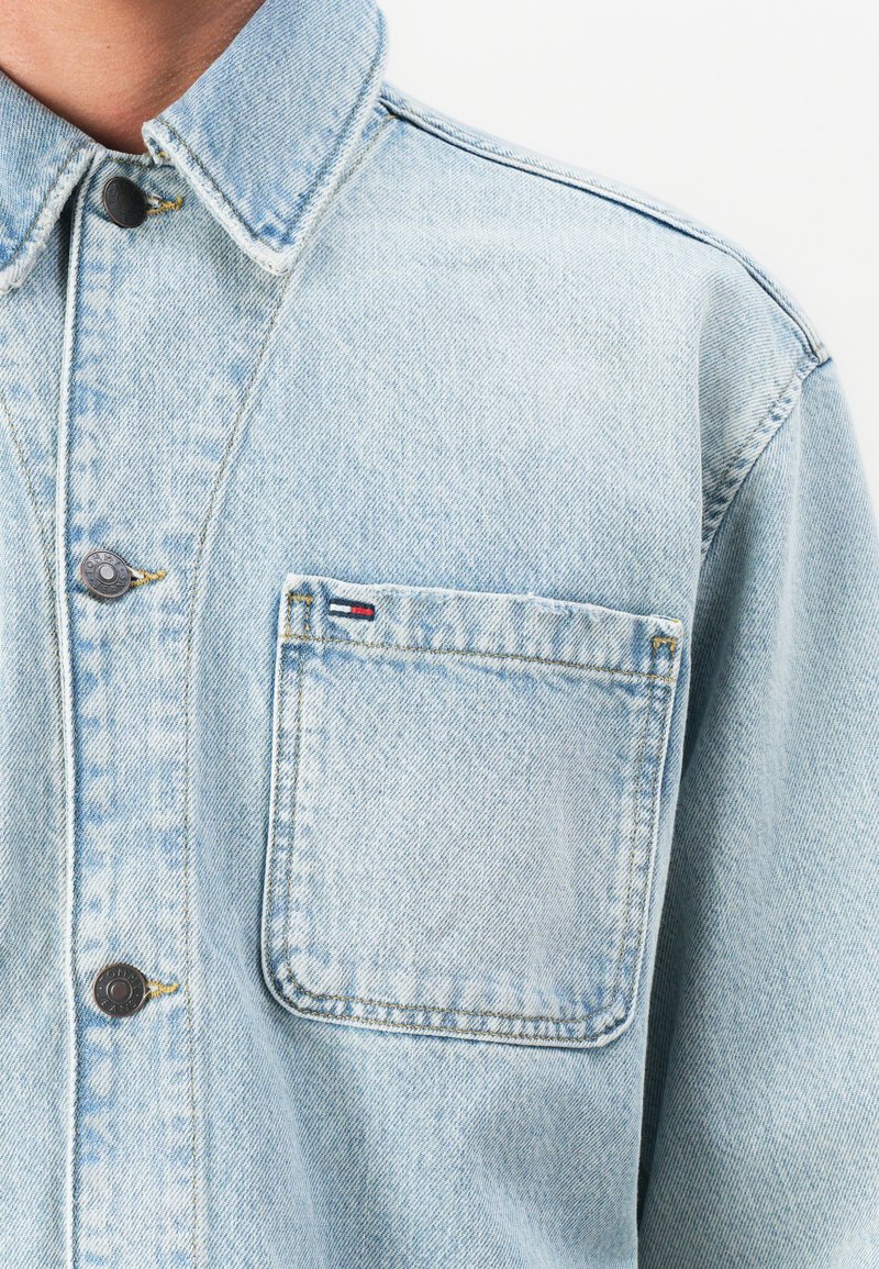 Veste en denim bleu clair avec des boutons en métal noir et une poche avant carrée présentant une petite étiquette logo rouge, blanche et bleue.
