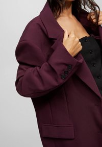 Vinröd kavaj med framträdande slag, strukturerade axlar och fyra svarta knappar som detaljer på ärmsluten. Har en sidoficka och en mjuk textur.