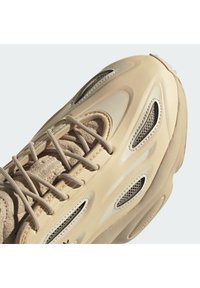 Beige sneakers med en texturerad läderoverdel, släta plastdetaljer, nätventilationshål och en vadderad sula. Snören i en ljus nyans.