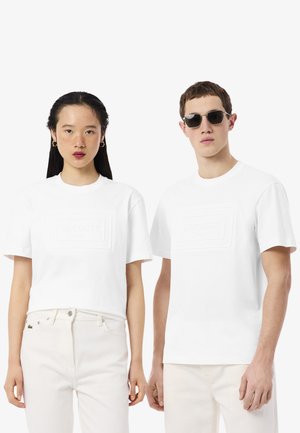 Dos modelos llevan camisetas blancas de manga corta con el logotipo en relieve de Lacoste y pantalones blancos; una modelo lleva pendientes dorados y la otra gafas de sol.