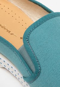 Teal canvas slippers met een gestructureerde bovenkant, contrasterend grijs stoffen paneel en geperforeerde beige binnenzool; geaccentueerd met witte stiksels.