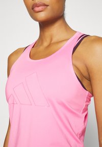 Vaaleanpunainen urheilutoppi, jossa on hieman kiiltävä pinta, pyöreä pääntie ja Adidas-logo. Ohuet mustat hihnat näkyvät alla.