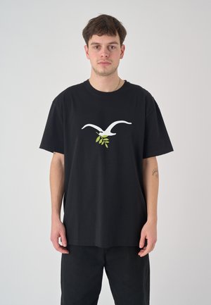 Junger Mann trägt ein lockeres schwarzes T-Shirt mit einem weißen Vogel und grünem Olivenzweig als Motiv, steht vor einem schlichten hellen Hintergrund.