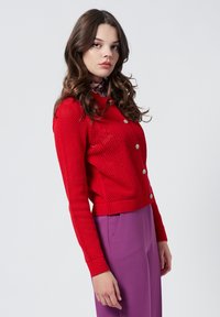 Cardigan rosso lavorato a maglia con un motivo testurizzato, scollatura rotonda e bottoni argentati. Abbinato a pantaloni viola a vita alta.