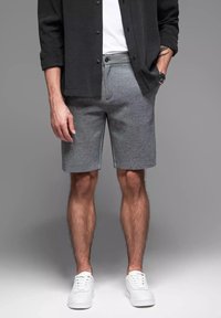 Graue Baumwollmischungsshorts mit figurnahem Bund, Seiten- und sichtbaren Nahtdetails. Kombiniert mit einem schwarzen Hemd mit Knöpfen und weißen Sneakers.