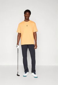 Oranje katoenen t-shirt met een klein zwart logo, gecombineerd met donkerblauwe broek en witte golfschoenen, met een golfclub in de hand en een handschoen aan.
