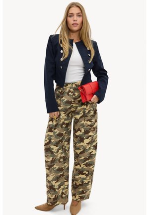Femme portant une veste courte bleu marine, un haut blanc, un pantalon large camouflage, des talons marron, tenant une pochette matelassée rouge.