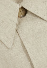 Camisa de lino beige con cuello puntiagudo y un botón metálico redondo con un diseño en relieve. La textura es ligeramente áspera y tejida.