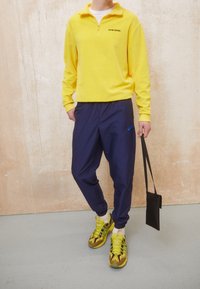 Żółta polarowa bluza na zamek, granatowe elastyczne spodnie z logo Nike oraz kolorowe buty sportowe o odważnym wzorze. Trzyma małą czarną torbę.