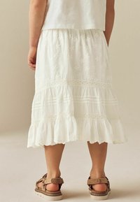 Jupe blanche à volants avec broderies florales et accents en dentelle. Elle présente un ourlet festonné et est assortie à des sandales brunes.