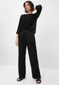 Pull en maille noir à tissage lâche et respirant ; associé à un pantalon large noir sur mesure. Les chaussures sont des sandales noires à enfiler.