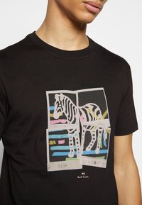 Černé bavlněné tričko s grafikou zebry v tlumených barvách, s abstraktními pruhy a bloky, a podpisem na spodní části.