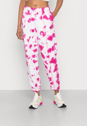 Kvinde iført hvide og lyserøde tie-dye joggingbukser med sort Nike-logo, hvide og lyserøde sneakers, stående mod en ensfarvet lys baggrund.