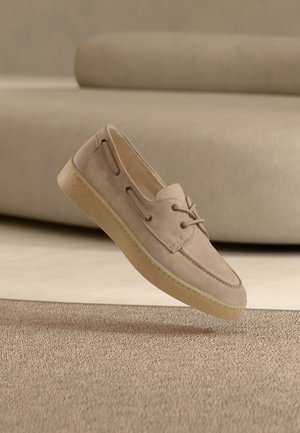 Beige suède schoen met een afgeronde neus, veters en zijopeningen. De zool is crèmekleurig met een gestructureerde afwerking. Zwevend boven een lichte ondergrond.