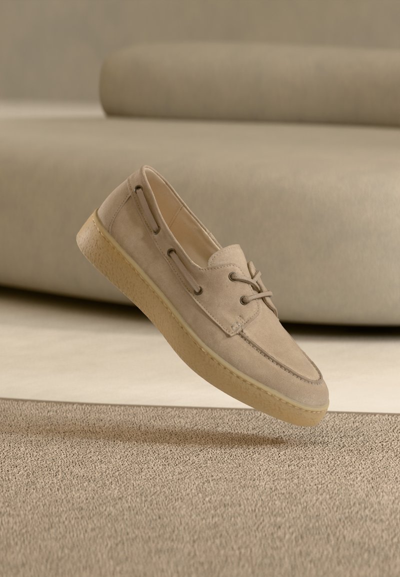 Μπεζ σουέντ παπούτσι με στρογγυλερή μύτη, κορδόνια και πλαϊνές περικοπές. Η σόλα έχει εκρού χρώμα με υφή. Επιπλέει πάνω από μια φωτεινή επιφάνεια.