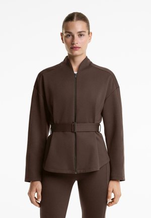 Vrouw in een getailleerde bruine ritsjack met een ceintuur in de taille en bijpassende bruine broek, staand tegen een witte achtergrond.
