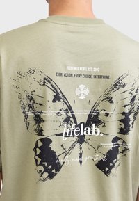 Olijfgroen katoenen T-shirt met een grote zwarte vlinderafbeelding op de achterkant, vergezeld van witte tekst en accenten bij het ontwerp.