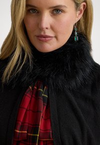 Guler din blană artificială de culoare neagră, cu o eșarfă cu model carouri roșii și negre vizibilă dedesubt, purtată peste o ținută închisă la culoare. Cercei detaliați cu accente verzi.