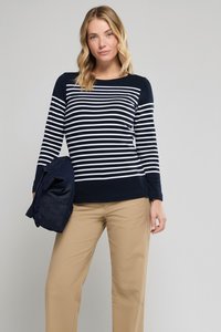 Frau mit blonden Haaren, die ein navy-blau-weiß gestreiftes Langarmshirt und beige Hosen trägt, hält in einer Hand eine dunkelblaue Jacke.