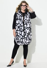 Svart och vit uppblåsbar väst med stora, djärva bokstäver, med dragkedja och sidofickor, lager på lager över en svart topp och tights.