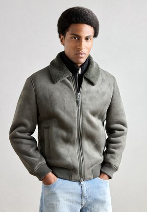 Olive green bomber jacket made of suede, featuring a textured fur collar, ribbed cuffs, and a zip closure. Worn over a black sweater and jeans.