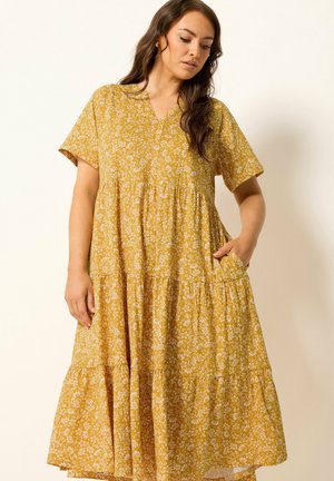 Kobieta w luźnej żółtej sukience w kwiaty z krótkimi rękawami, stojąca i patrząca w dół, jedna ręka w kieszeni, na tle jednolitego jasnego tła.