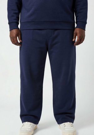Uomo che indossa pantaloni della tuta blu navy e felpa abbinata con sneaker bianche, in piedi contro uno sfondo chiaro e uniforme.