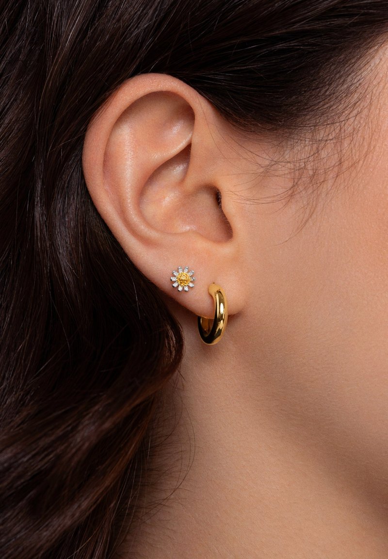 Pendiente de aro dorado y un pendiente en forma de flor con piedras claras y amarillas. Superficie de metal suave con acabado pulido. Primer plano de una oreja.