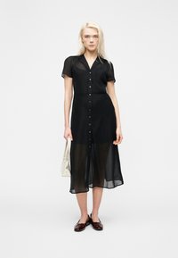 Γυναίκα με μακριά ξανθά μαλλιά που φοράει διαφανές μαύρο μίντι φόρεμα με κουμπιά μπροστά, κρατώντας ένα λευκό τσαντάκι, και στέκεται μπροστά σε απλό φόντο.