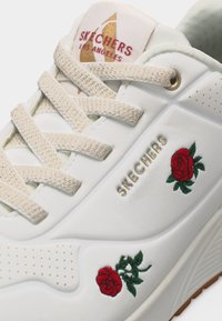 Biały sneaker Skechers z beżowymi sznurowadłami, złotym logo Skechers oraz haftowanymi czerwonymi różami z zielonymi liśćmi po bokach.