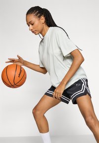 Chemise vert clair à manches courtes associée à un short de sport noir avec des rayures blanches. Tient un ballon de basket orange, concentré sur le ballon.
