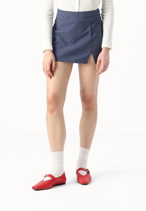 Vrouw met een blauwe geruite minirok, witte top met lange mouwen, witte sokken en rode Mary Jane platte schoenen, staand tegen een witte achtergrond.