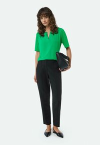 Green short-sleeve blouse with a notch collar, paired with fitted black trousers and pointed black flats, holding a black bag.