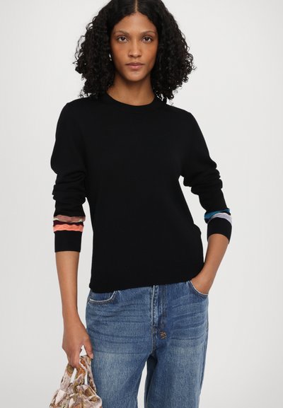 Svart strikkegenser med lange ermer som har flerfargede bølgende detaljer ved mansjettene. Sammen med denimjeans og en teksturert veske.