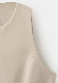 Vêtement beige sans manches avec un col rond et une texture lisse, montré depuis l'épaule et la partie supérieure de la poitrine.
