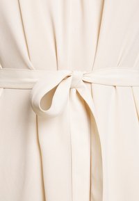 Ceinture en tissu crème nouée en un simple nœud autour de la taille d’un vêtement crème assorti avec des plis doux.
