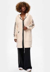 Vrouw met krullend haar, gekleed in een oversized beige ritssluiting hoodie en zwarte wijde broek met witte sneakers, staand tegen een effen achtergrond.